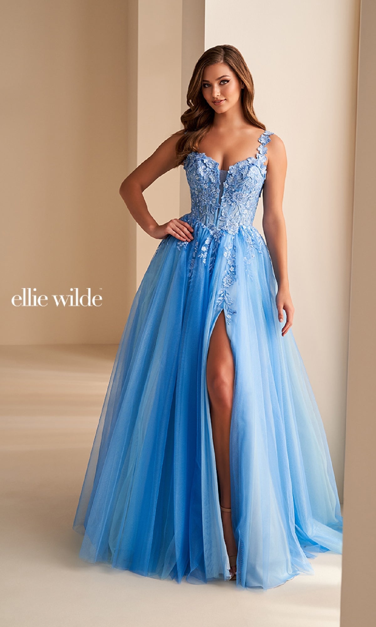 A woman indoors wears the Ellie Wilde EW36214, a light blue sleeveless floor-length A-line tulle prom dress with floral lace details and a thigh-high slit.